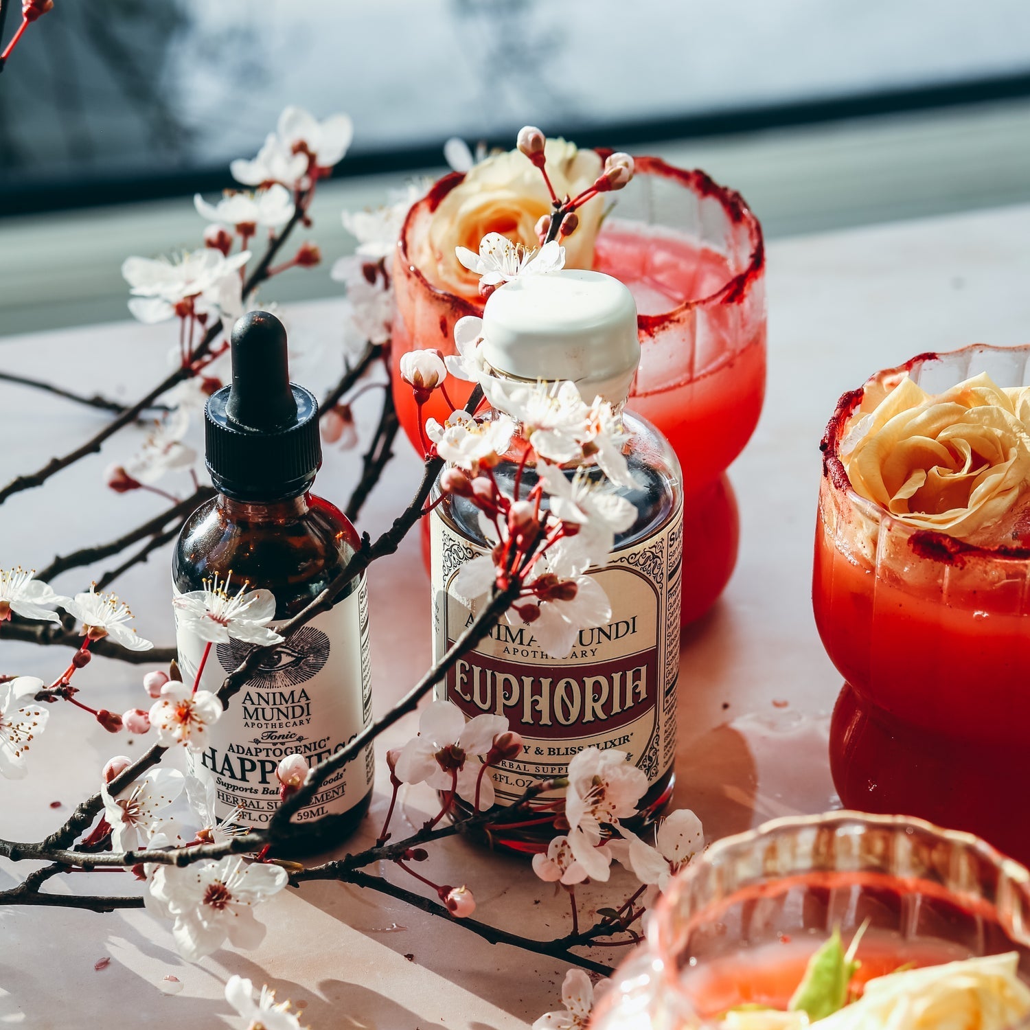 Glasses of red cocktails with floral garnishes, bottles of Euphoria and another brand, and orange slices on a light surface.