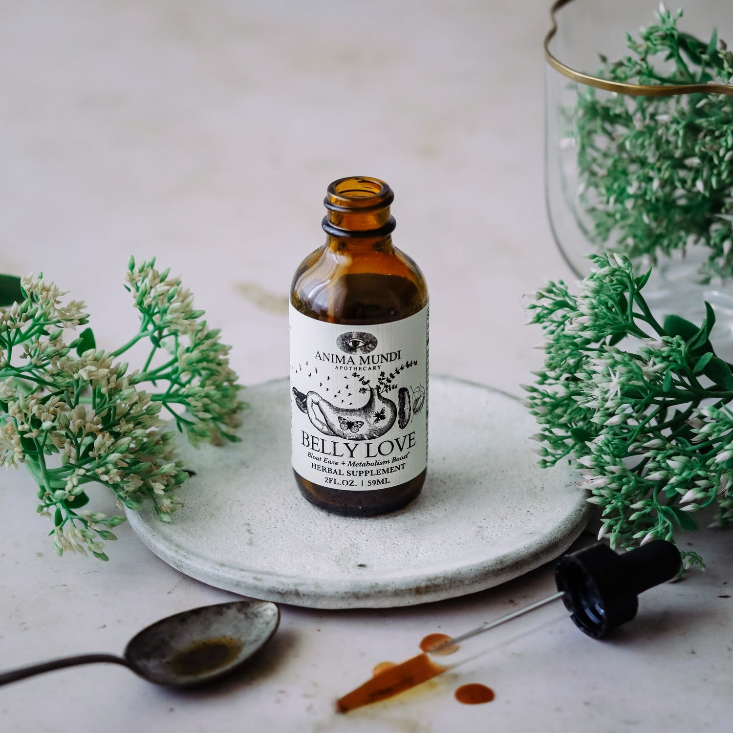 BELLY LOVE Tonic bottle surrounded by herbs and a spoon on a stone plate.