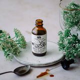 BELLY LOVE Tonic bottle surrounded by herbs and a spoon on a stone plate.