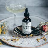 Bottle of 'Anima Mundi Relax' supplement on a decorative plate with a glass of white wine and flowers.