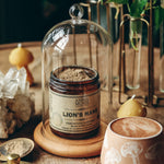 Ceramic container labeled 'Lion's Mane' on a wooden surface with a glass dome, mug, and decorative elements.