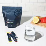 Pure Hydration drink mix sachets with a glass of water, lemon, and watermelon on a kitchen counter.