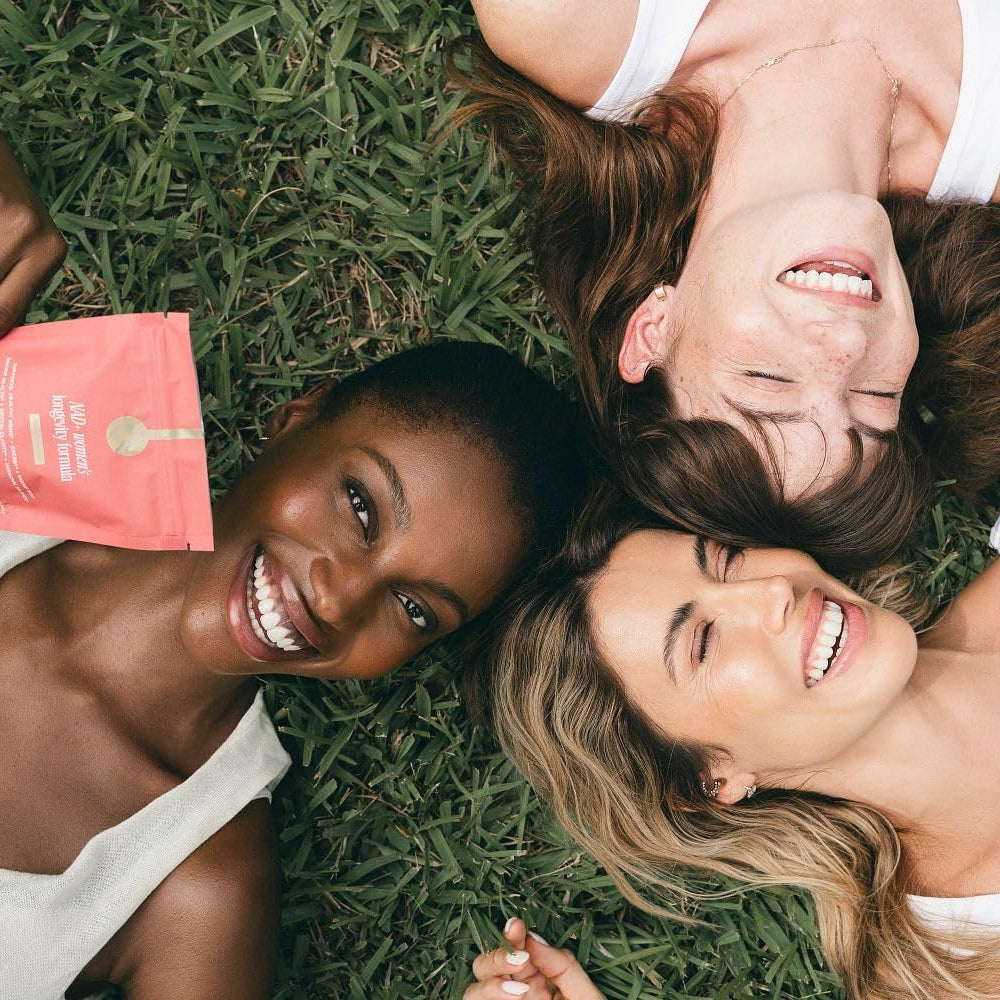 Three women lying on grass with a book, smiling.