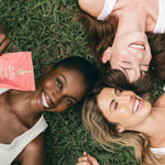 Three women lying on grass with a book, smiling.