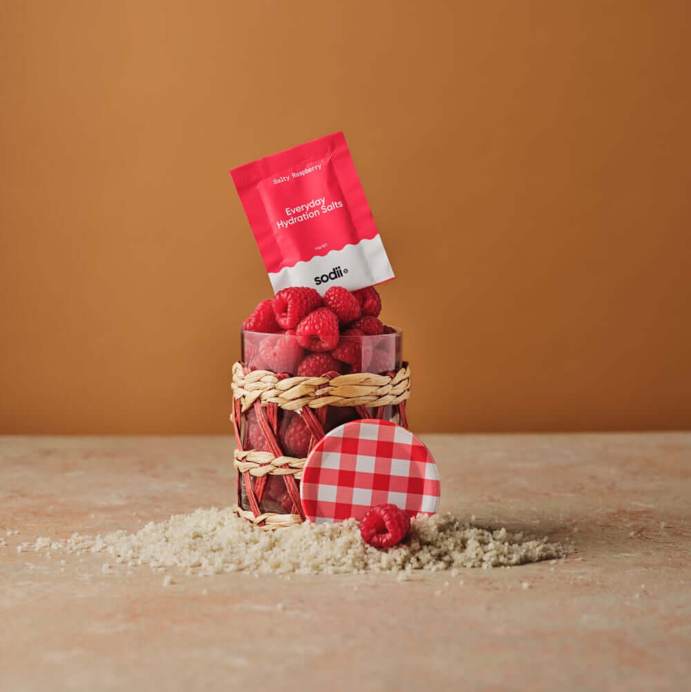 Everyday Hydration Salts Raspberry package displayed with raspberries in a basket on a sandy surface.